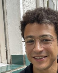 Smiling, curly dark hair and round wire-rimmed glasses, outside a cafe.